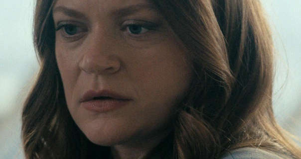 Colby Minifie is seen looking slightly downwards while in deep thought. She sits in front of a large skyscraper window during the day. Colby has her light brown hair loose and partly curled. She wears a matt foundation with mascara and a dark pink satin lipstick.