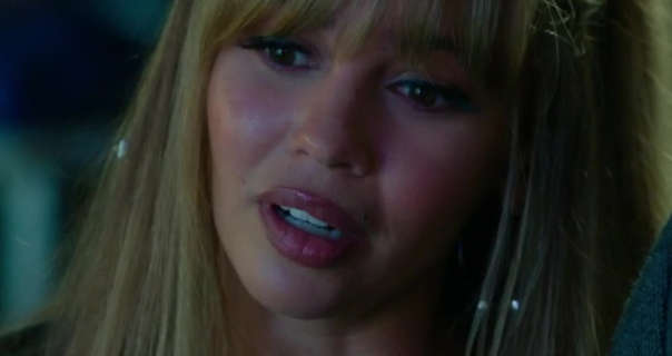 Vanessa Morgan is seen looking thoughtful as she glances downwards. She wears her thick long hair coloured light blonde, straightened and loose. Vanessa appears in blush, mascara, matt foundation and a dark red lipstick.