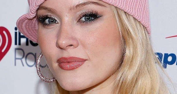 Zara Larsson wears a light pink knitted beanie with a pair of gemstone large hoop earrings. She is seen in a soft blush, black mascara and a dark red lipstick. Zara stands in front of a dark white event wall with several logos across it.