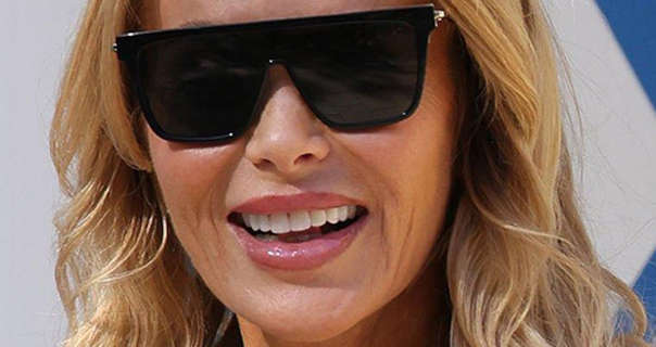 Amanda Holden is seen in a pair of large black, flat top sunglasses. She walks past a long blue and white sign. Amanda wears her blonde hair loose and styled in waves. She stars in a light foundation and a dark pink gloss lipstick.