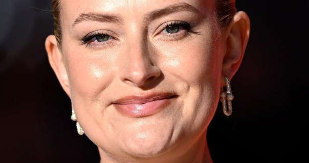Amelia Dimoldenberg is seen outdoors at a nighttime event with darkness around her. She wears her blonde hair tied back in a slick bun. Amelia wears a high coverage foundation with black mascara, hanging gemstone studded earrings and a dark pink lipstick.
