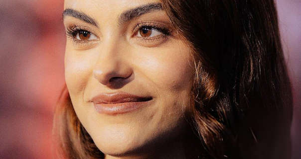 Camila Mendes is seen in a matt foundation, mascara and a dark pink soft shine lipstick. She wears her black hair loose, tinted with highlights and curled. Camila stands in front of a purple, pink and orange hue poster wall at an event.
