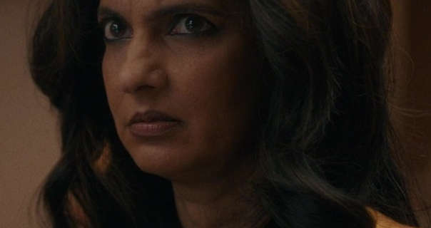 Poorna Jagannathan frowns slightly with an agressive look. She wears her thick black hair loose and in soft waves. Poorna appears in a dark room that has wooden display shelves and beige walls. She wears foundation with black eyeliner, mascara and a deep pink lipstick.