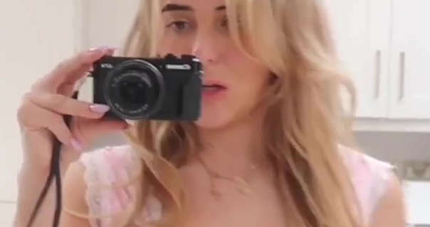 Em Sheldon is seen in her white marble kitchen at her home in London. She has her long blonde hair loose and in natural curls. Em holds her black Canon camera in one hand as she films. She wears a light pink dress with pink nails, light foundation, styled brows and a pink lipstick.