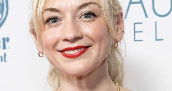 Emily Kinney wears a red lipstick with mascara, small gold hoop earrings, light foundation and a gold necklace. She stands in front of a white event wall that has dark teal writing and logos across it. Emily has her long blonde hair and tied back.