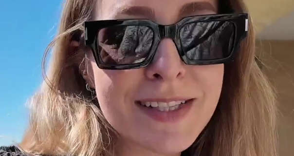 Fleur Bell walks past a row of shops outdoors in the sunshine in Florence. She wears her long thick blonde hair loose and styled naturally. Fleur appears in a pair of large black frame sunglasses, gold earrings, matt foundation and a dark pink lipstick.
