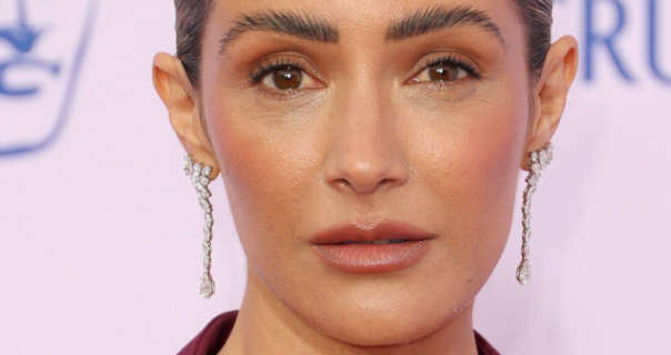 Frankie Bridge wears her hair tied up in a tight bun. She is seen in a matt foundation, feathered brows, blush, dark pink lipstick and a pair of hanging diamond studded earrings. Frankie stands in front of a white event wall that has think blue text and blue logos across it.