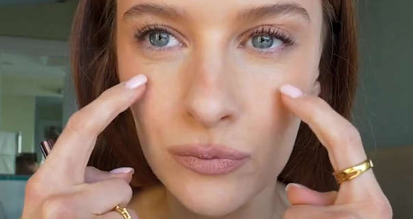 Victoria Magrath points to bags under her eyes with the index fingers of both her hands. She wears her long hair loose, down and pushed back. Victoria sits in a hotel bedroom. She appears in a matt foundation, gold rings, curled lashes, blush and a dark pink lipstick.