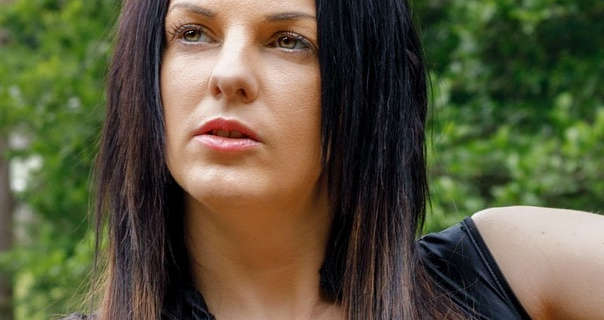 Izzy Rose looks upwards and to one side as she poses outdoors in front thick green bushes and trees. She has her long black hair loose, straightened and with blonde tips. Isabella wears a matt foundation with curled lashes and a light pink gloss lipstick.