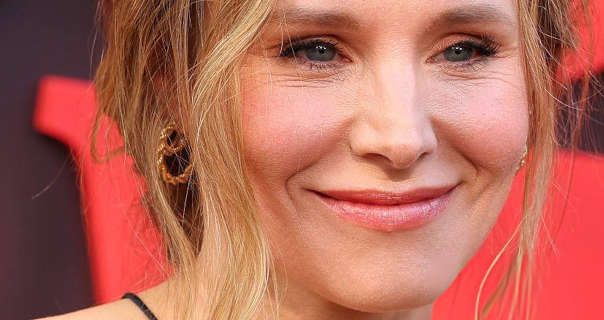 Kristen Bell is seen in front of a black wall with chunky pastel red 3D writing on it. She wears her long blonde hair tied back with multiple strands pulled around her face. Kristen stars in a soft foundation, gold textured earrings, mascara, blush and a light pink lipstick.
