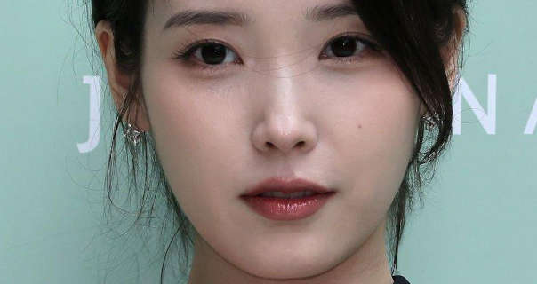 Lee Ji-eun stands in front of a mint colour event wall that has white text on it. She wears her long black hair tied up and back. Lee Ji-eun appears in a soft foundation, mascara, silver gemstone earrings and a red lipstick.