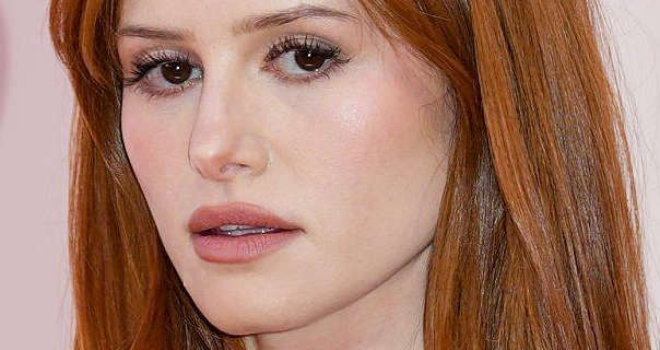 Madelaine Petsch appears in front of a dark white wall at an event. She wears her red hair straightened and loose. Madelaine looks into the camera with a natural expression. She wears a matt foundation with black mascara, light blush and a pink lipstick.