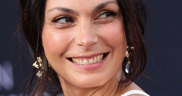 Morena Baccarin smiles and looks to the side. She is standing in front of a black event wall with out of focus white text on it. Morena has her long hair tied back and up with some strands around her face. She wears a white strappy dress with hanging gold with glass earrings, light foundation, blush, mascara and a baby pink lipstick.
