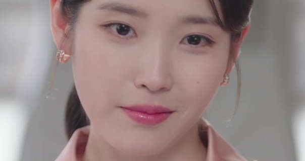 Lee Ji-eun smirks a little. She sits in a large light and airy open plan room during a meeting. Lee Ji-eun wears her long black hair tied back in a ponytail. She stars in a smoothing foundation, gold gemstone hoop earrings and a pink lipstick.