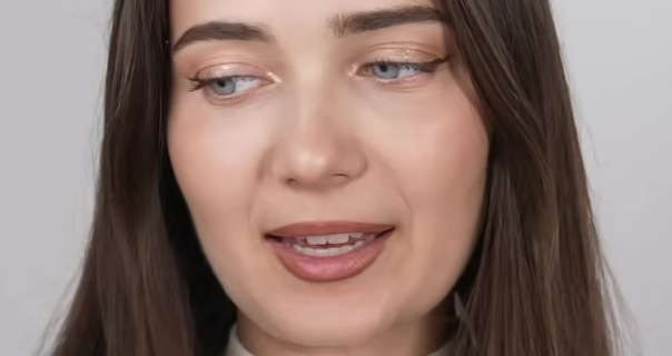 Roxxsaurus is seen in front of a dark white wall. She has her long brown hair straightened and loose. Roxi wears a matt foundation with subtle blush, black eyeliner, light eye shadow and a dark red overlined lipstick. She looks slightly downwards and to one side as she talks.