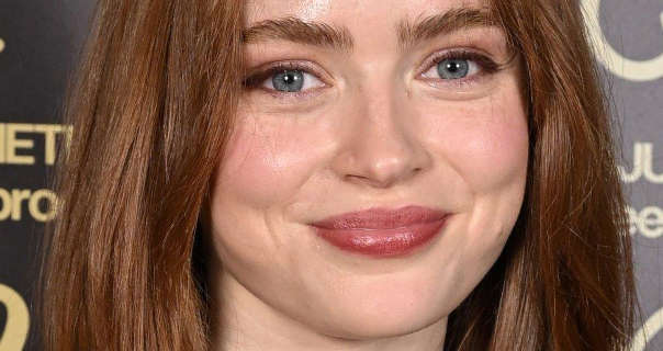 Sadie Sink is seen smiling as she stands in front of a grey event wall that has yellow gold writing on it. She wears her long red hair straightened and loose. Sadie appears in a matt foundation, blush, curled lashes and a red shiny lipstick.