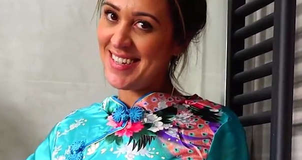 Samantha smiles as she stands in a cream tile bathroom next to a long black heated towel rack. She has her long hair tied up and back in a bun. Samantha wears a natural foundation with mascara, pink lipstick and a sea green cheongsam.