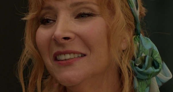 Lisa Kudrow smiles and looks to the side. She appears in front of a dark grey stage wall. Lisa has her ginger colour hair tied back with some wavy strands pulled out to frame her face. Lisa wears a matt foundation with black mascara, pink lipstick and blue green headscarf.
