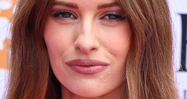 Victoria Magrath is seen in front of a dark white event wall that has orange and black text and logos on it. Victoria has her long hair loose and coloured with brown and blonde highlights. She wears a matt foundation with mascara, blush and a pink satin lipstick.