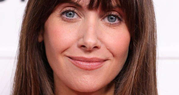 Alison Brie stars in front of a dark white event wall that has black text and logos across it. Alison has her long brown hair loose, straightened and with a full fringe. She wears a light coverage foundation with mascara, blush and a nude sheen lipstick.