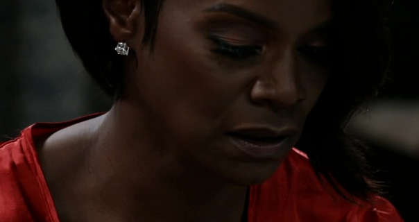 Rashan Ali looks down while sitting on a sofa in a dark living room at night. She wears her short black hair loose and curled inwards. Rashan wears a red robe with foundation, dark mauve lipstick, eyelash extensions and a pair of diamond earrings.