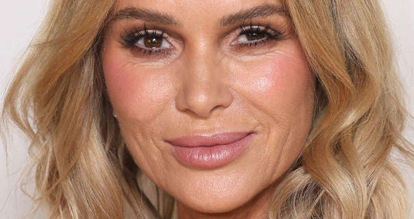 Amanda Holden is seen with her long blonde hair loose and in layered waves. She stands in front of a dark white wall. Amanda wears heavy blush with a dewy foundation, mascara and a light pink lipstick.