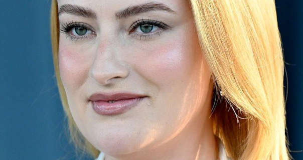 Amelia Dimoldenberg is seen in front of a light and dark blue curtain wall with bright sunlight hitting the side of her long blonde hair. She wears a light pink blush with matt foundation, curling mascara, pink mauve gloss lipstick and a pair of gemstone earrings.
