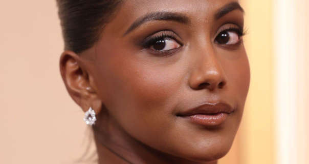 Charithra Chandran turns her head slightly to look directly into the camera. She stands in front of a pale yellow and orange hue wall. Charithra has her black hair tied back and up. She wears a soft blush with foundation, mascara, dark pink gloss lipstick and a pair of diamond studded earrings.