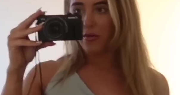 Em Sheldon films herself through a hotel room mirror while on holiday in Greece. She holds her black Canon camera in one hand. Em is seen with her long, thick blonde hair straightened and loose. She wears foundation with a dark pink lipstick and a light blue one shoulder dress.