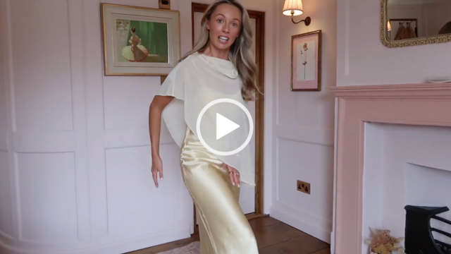 Josie Irons stands near the doorway of her white and dusty pink wood panel living room at her home. She is seen in a long yellow dress and a sheer yellow cape covering. Josie has her thick blonde hair loose and in gentle waves. She appears in a dewy foundation, pink lipstick and several gold rings.
