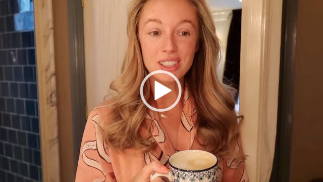 Josie Irons is seen in the bathroom area of a guest room at Estelle Manor in the early morning. She appears in front of a white wood door frame with a natural wood and blue tile shower area to one side. Josie wears her long blonde hair loose and in soft waves. She holds a blue and white cup in her hands and wears no makeup.