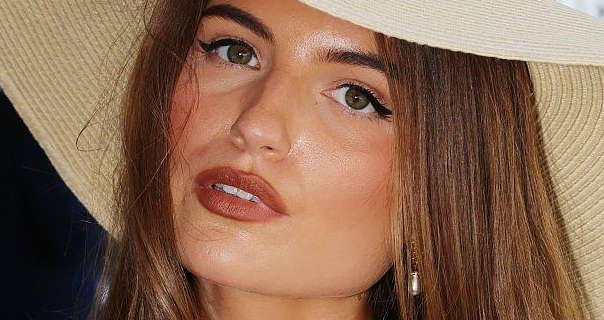 Grace Foley leans her head to one side as she stands in a dark event seating area. She wears a light woven wide brim hat. Grace has her long brown hair loose and with highlight tints. She appears in a dewy foundation, black eyeliner, mascara, dark pink lipstick and a pair of hanging pendant earrings.