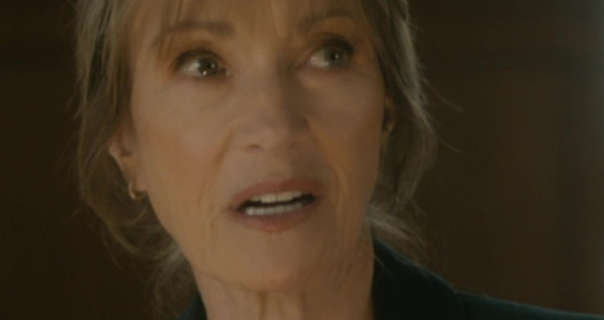 Jane Seymour is seen in front of a brown wall. She glances up and to one side. Jane has her long hair tied back in a bun. She wears a soft foundation with mascara, pink lipstick and a pair of small gold hoop earrings.