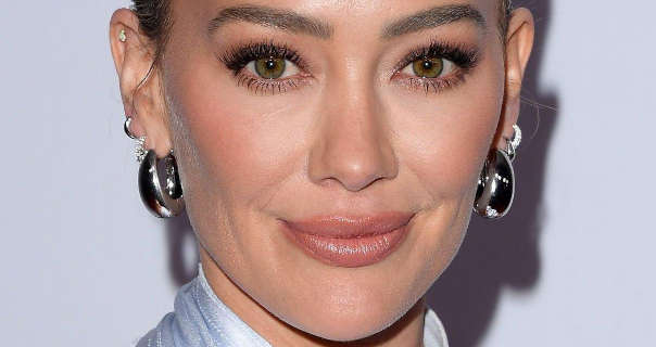 Hilary Duff stands in front of a dark white wall at an event. She has her long blonde hair tied back and up in a tight bun. Hilar wears a pair of large and chunky hoop earrings with a few smaller silver rings and studs. She stars in a matt foundation, pink blush, mascara and a dark pink lipstick.