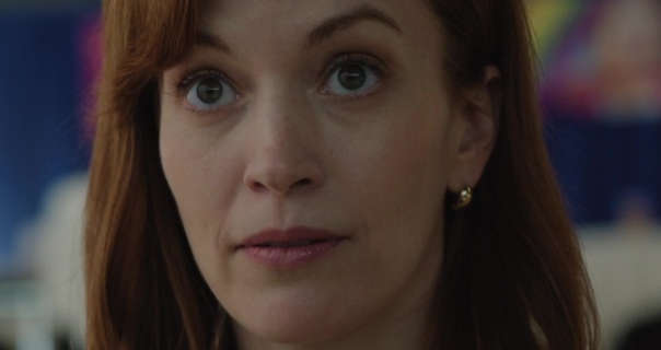 Britt Lower sits in a small auditorium with a blue wall and colourful abstract paintings behind her. She has her dark red hair loose, straightened and with a side parting. She wears gold earrings with a matt foundation, mascara, light blush and a dark pink sheen lipstick.