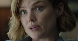 Erin Richards is seen with her short blonde hair in gentle waves. She sits in a large open plan kitchen area in a flat light. Erin wears a soft foundation with curled lashes, light blush, pale pink lipstick and a pair of small gold hoop earrings.