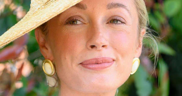 Josie Irons looks to the side and smiles. She stands in front of a green and red leafy bush outside in the sunshine. Josie wears a woven hat at an angle with her long blonde hair tied back in a bun. Josie stars in a soft foundation, light pink blush, mascara, dark pink lipstick and a pair of large gold and white circular earrings.
