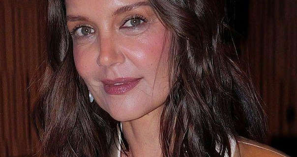 Katie Holmes stands in front of a dark wood effect wall. She wears her long black hair layered and down. Katie wears a gold blazer with hanging earrings, blush, mascara and a deep red lipstick.