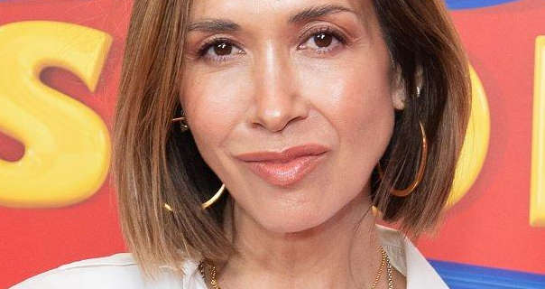 Myleene Klass stands in front of a yellow, red and blue movie poster wall. She has her light brown hair cut to shoulder length and straightened. Myleene wears a matt foundation with mascara, pink lipstick, two dainty chain necklaces and a pair of large thin gold hoop earrings