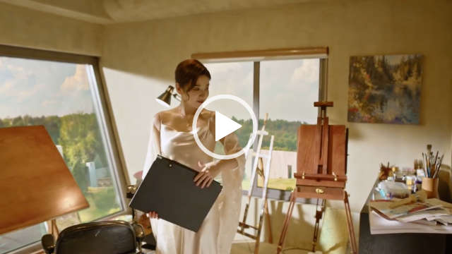 Huang Yi stands in a painting room surrounded by easels and art equipment. She appears in front of light stone walls and modern large windows that look out to grass lawns and a forest of trees. Huang Yi wears a white shawl over a long white cowl neck dress. She has her hair tied back with strands pulled out around her face.