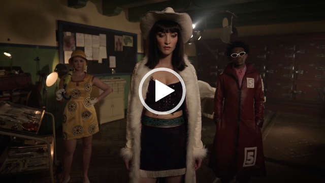 Jazzara Jaslyn stands to one side in a large dark cream, green and brown autopsy room. She wears a yellow wheel print dress with a wide brim yellow had. Jazzara holds a closed umbrella over her shoulder with one hand with her other hand on her hip. Her short blonde hair is straightened and cut at a sharp angle.