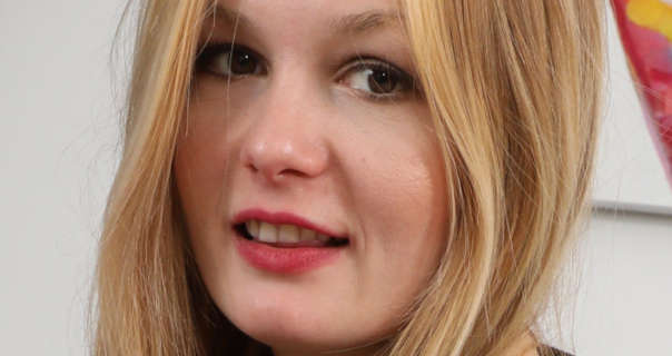 Freya H turns and looks into the camera over a shoulders. She appears in front of a dark white wall with a large abstract brush stroke painting hung on it. Freya has her long blonde mix hair loose and naturally styled. She wears a soft foundation with mascara and a dark red lipstick.