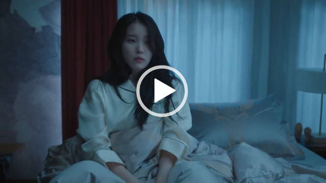 IU sits up in bed during the night looking sad. She has her long black hair loose. IU wears a white and black trim pair of pyjamas. She is seen on a large light grey bed with sheer white and deep red curtains behind her.