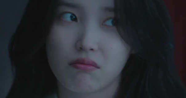 IU is seen in a large dark bedroom. She looks to one side unhappily. IU wears a soft foundation with a light pink lipstick and no jewellery. She has her long thick black hair loose in a natural style.