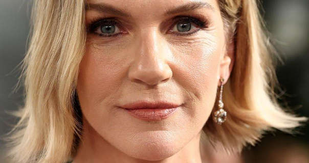 Rhea Seehorn is seen against an out of focus light grey event wall with a darker entrance area to one side. She wears her blonde hair at shoulder length and slightly wavy. Rhea wears a pair of pendant earrings with a dewy foundation, mascara and a dark pink lipstick.