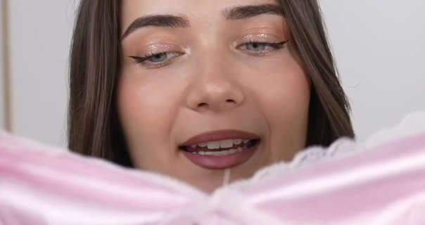 Roxxsaurus holds up a light pink dress bought through an Instagram advert. She has her long thin brown hair loose and straight. Roxi sits in front of a dark white wall with a wood frame painting to one side. She wears a matt foundation with blush, winged eyeliner, pale glitter eye shadow and a dark red matt lipstick.