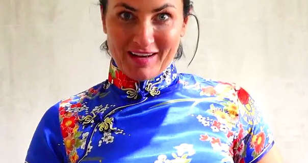 Sarah Wild talks to the camera while standing in front of a cream and light grey bathroom tiled wall. She has her long black hair tied back and up in a bun. Sara wears a blue floral print cheongsam, natural foundation, mascara and light pink lipstick.