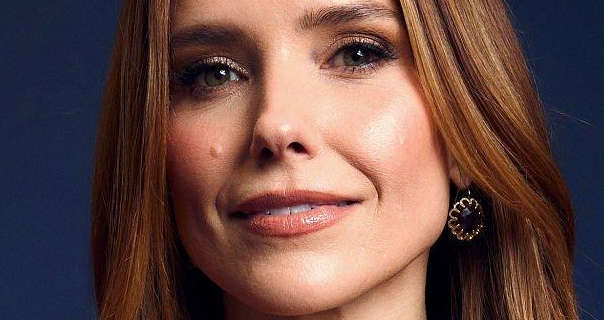 Sophia Bush is seen against a deep navy blue background. She has her long brown hair loose and with blonde highlights. Sophia stars in a matt foundation, black mascara, blush, dark pink lipstick and a pair of large handing gold and gemstone earrings.