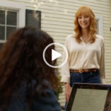Lisa Kudrow appears outdoors in partial sunlight. She stands in front of Abbi Jacobson who is sitting at a low table on a MacBook with her back to the camera. Lisa wears a long sleeve white blouse tucket into a pair of dark denim jeans. Her long light orange hair is loose and in curled with a long fringe.