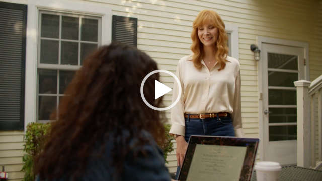 Lisa Kudrow appears outdoors in partial sunlight. She stands in front of Abbi Jacobson who is sitting at a low table on a MacBook with her back to the camera. Lisa wears a long sleeve white blouse tucket into a pair of dark denim jeans. Her long light orange hair is loose and in curled with a long fringe.